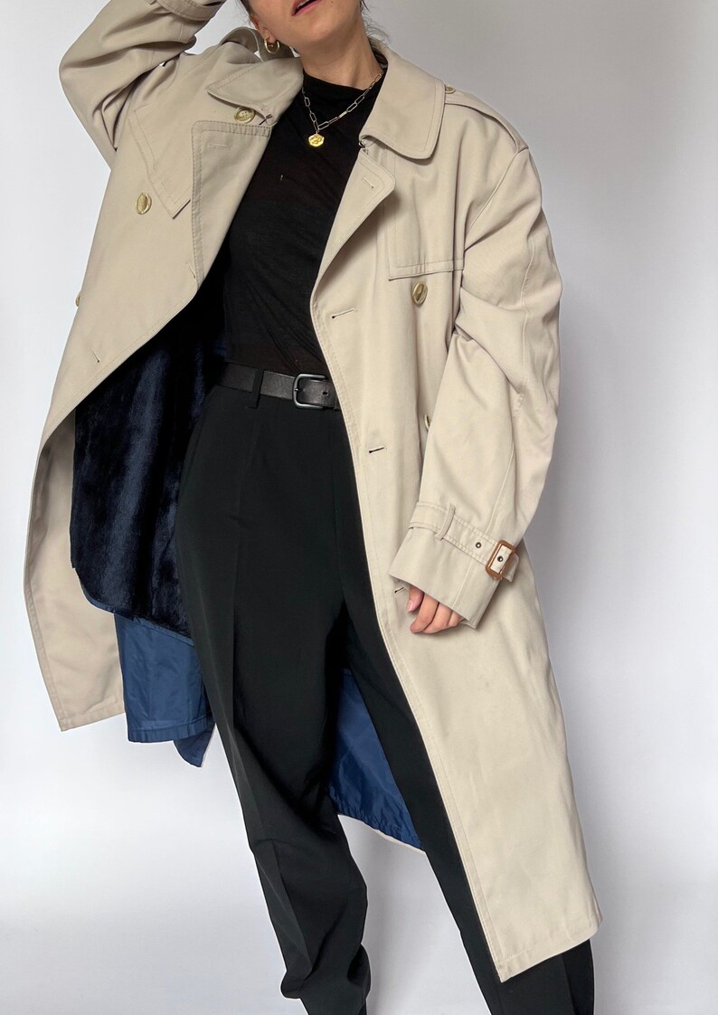 May include: A beige trench coat with a brown leather belt and a blue lining. The coat has a double-breasted closure and a collar.