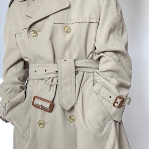 May include: A beige trench coat with a brown leather belt and brown buttons. The coat has a double-breasted closure and two flap pockets.