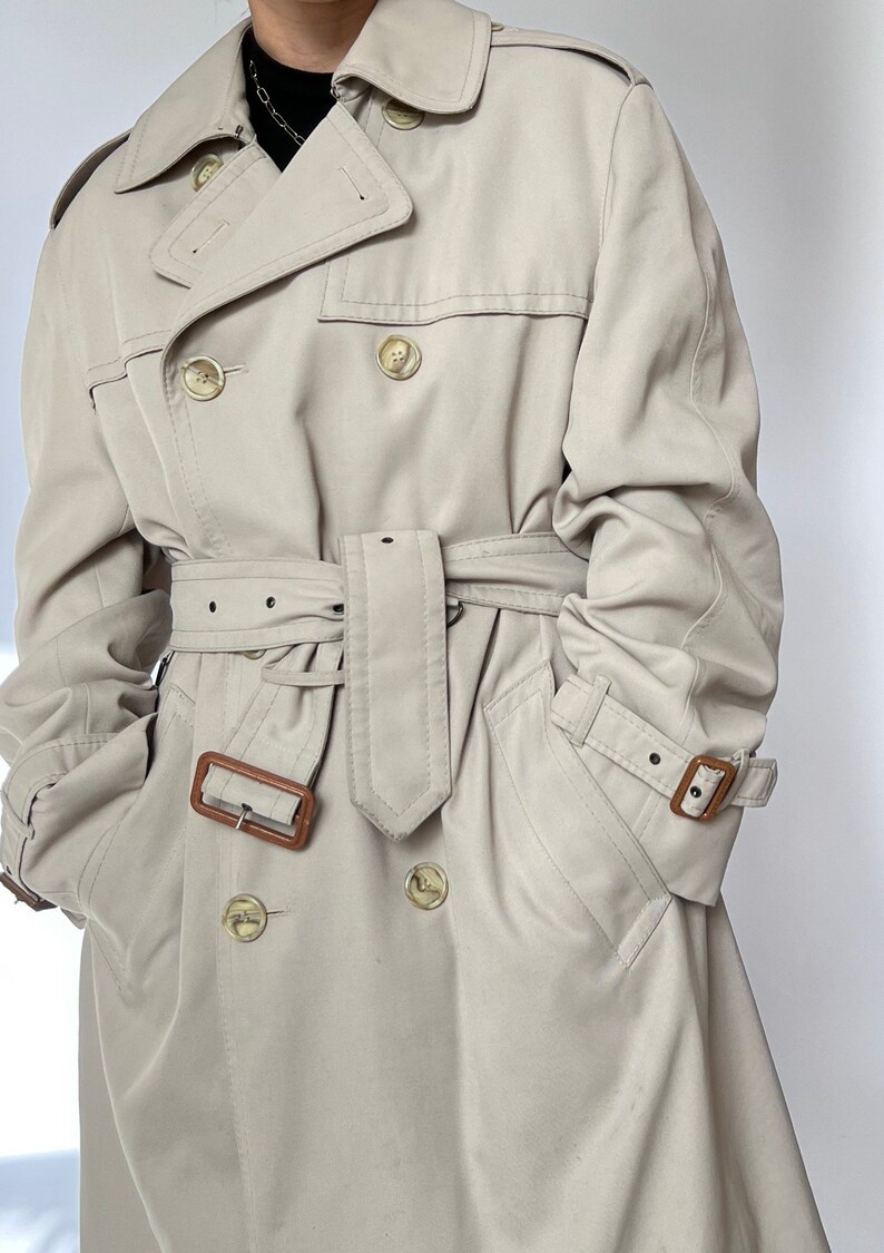 May include: A beige trench coat with a brown leather belt and brown buttons. The coat has a double-breasted closure and two flap pockets.