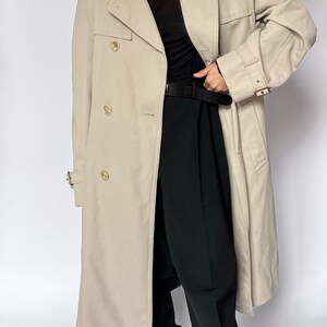 May include: A beige trench coat with a double breasted closure and a belt. The coat has a classic design and is made of a lightweight fabric.