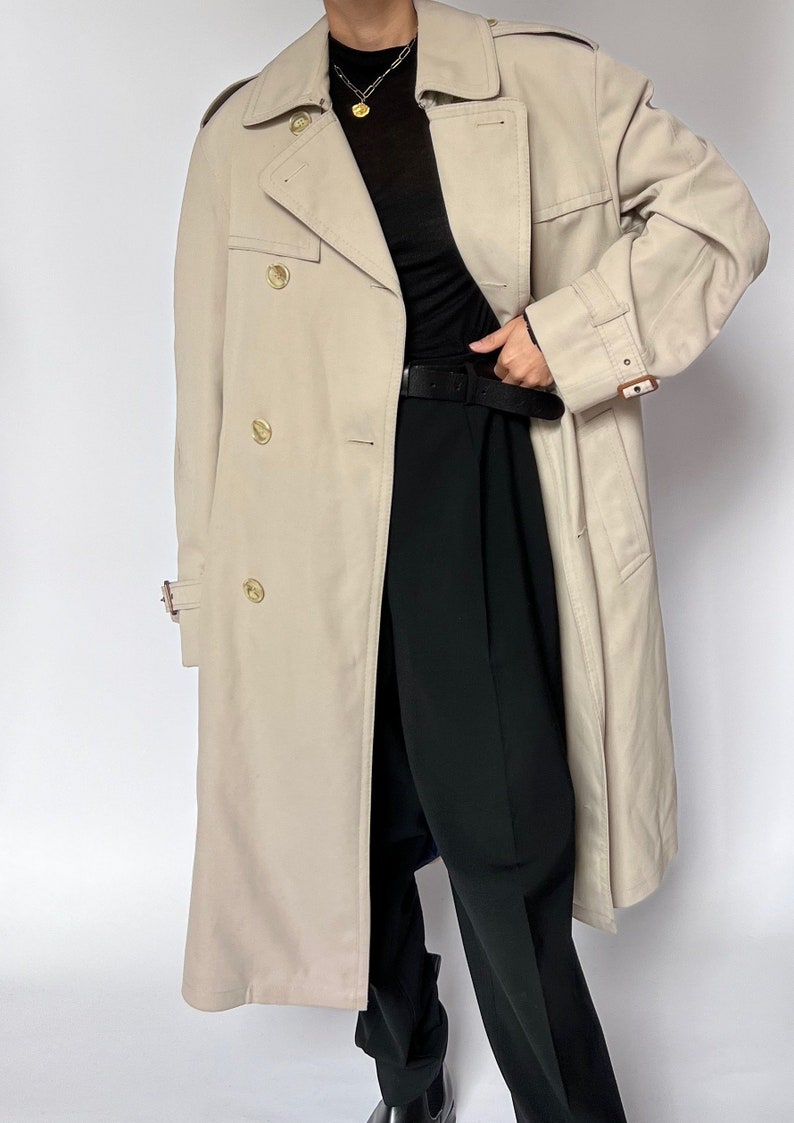 May include: A beige trench coat with a double breasted closure and a belt. The coat has a classic design and is made of a lightweight fabric.