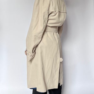 May include: A beige trench coat with a belt and a button closure. The coat has a classic design and is made of a lightweight fabric.