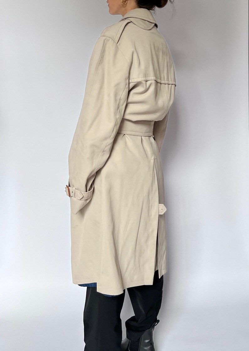 May include: A beige trench coat with a belt and a button closure. The coat has a classic design and is made of a lightweight fabric.