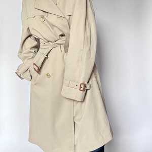May include: A beige trench coat with a brown leather belt and brown leather buckle accents. The coat has a double-breasted closure and a collar.