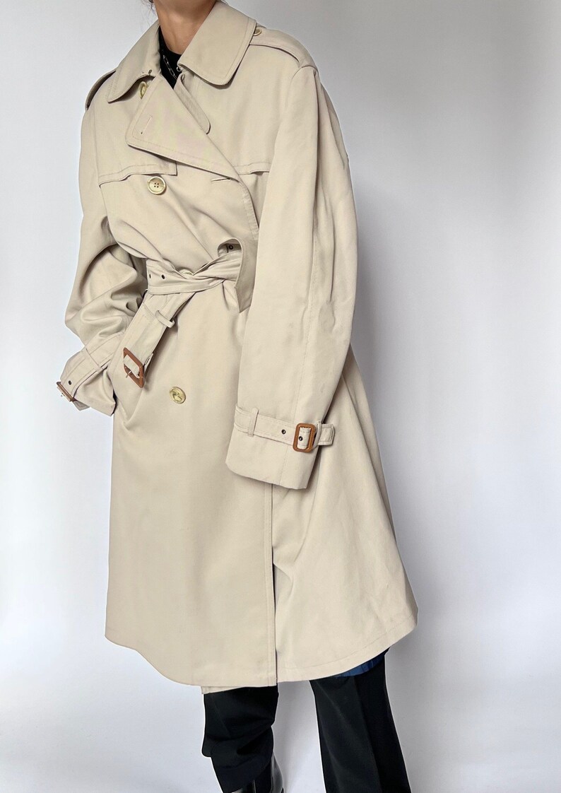 May include: A beige trench coat with a brown leather belt and brown leather buckle accents. The coat has a double-breasted closure and a collar.