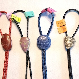 May include: Six different bolo ties with various colored stones and leather cords. The stones include red, purple, blue, and gray. The cords are red, black, and blue.