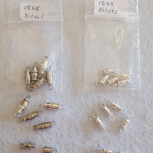 May include: Two clear plastic bags labeled "15x5 Silver" and "16x4 Silver" containing silver-colored magnetic clasps. Several clasps are scattered outside the bags. These clasps are used for jewelry making.