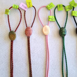 Könnte beinhalten: Fünf Bolo-Krawatten mit geflochtenen Kordeln in Rot, Braun, Pink, Grün und Schwarz. Jede Krawatte hat einen einzigartigen ovalen Stein- oder Muschelschieber, darunter Tigerauge, gesprenkelter Stein und Perlmuttschale, mit kleinen Papieretiketten.