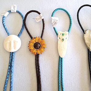 May include: Four bolo ties with different designs. One has a white moon face, another a sunflower, a third a carved ivory-colored insect, and the last a silver arrowhead. The ties have braided leather cords in blue, brown, teal, and black.
