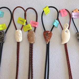 May include: A collection of bolo ties with various stone and carved pendants. The ties feature black or brown braided cords and are adorned with tags. The pendants include a variety of shapes and colors, showcasing natural textures and patterns.