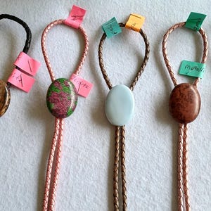 Könnte beinhalten: Fünf Bolo-Krawatten mit ovalen Cabochon-Steinen in verschiedenen Farben und Mustern, darunter Braun, Grün und Pink, Hellblau sowie gesprenkeltes Braun. Jeder Stein ist auf einer geflochtenen Kordel mit einem kleinen farbigen Papieretikett befestigt.