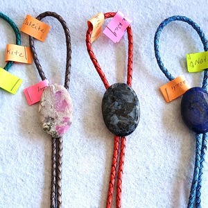 May include: Five bolo ties with different colored cords and stone pendants. The stones are turquoise, pink, black, blue, and white. The cords are green, brown, red, blue, and pink.
