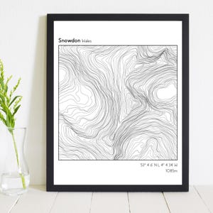 May include: A black and white topographic map print of Snowdon, Wales. The print shows contour lines and the coordinates 53° 4.6' NL 4° 4.34' W. The print also includes the text "Snowdon Wales" and "1085m".