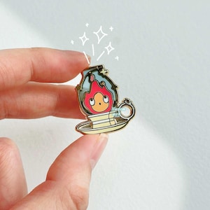 May include: Gold-tone enamel pin featuring a cartoon character with red hair and a yellow body, inside a clear glass dome with a blue and white design. The character is sitting on a yellow saucer with a gold rim. The pin has a gold-tone metal clasp on the back.