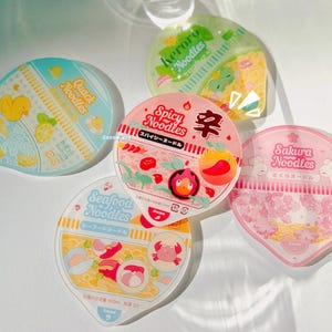 Könnte beinhalten: Draufsicht auf fünf Instant-Nudel-Behälter. Die Behälter weisen farbenfrohe Designs und Texte auf, darunter "Quack Noodles", "Keroro Noodles", "Spicy Noodles", "Sakura Noodles" und "Seafood Noodles". Jeder Behälter hat eine einzigartige Illustration, wie eine Ente, ein Frosch und eine Krabbe.