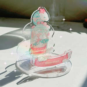May include: A clear acrylic stand featuring a pink and blue cartoon character holding a bottle. The bottle has a pink and blue label with Japanese text that reads "守護ーGuardian". The stand has a pink banner that reads "Guardian".