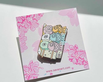 May include: Enamel pin featuring colorful Japanese sweets, including fish-shaped taiyaki and mochi on a rectangular tray. The pin is set on a card with pink cherry blossom illustrations and the website address www.savenart.com.