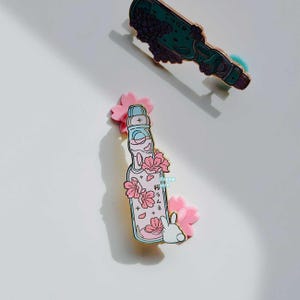 May include: Two enamel pins featuring Japanese soda bottle designs. One is green with purple floral accents, the other is pink with cherry blossoms and a white rabbit. The pins are on a white background.