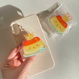May include: A clear acrylic charm featuring a cartoon pudding with a golden crown. The pudding is yellow and orange, with a small plate and the text "Seven art". The charm is attached to a phone case.