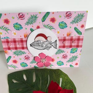 May include: A handmade gift box with a pink floral and tropical leaf pattern. A circular emblem features a fish wearing a Santa hat. A red and white checkered ribbon accents the box. A pink flower and a green leaf with a red poinsettia are in front.