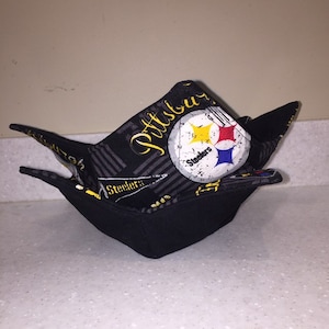 Op de afbeelding: Een zwarte stoffen kom met een Pittsburgh Steelers ontwerp. De kom is opgevouwen en heeft een zwart-grijs gestreept patroon met de woorden "Pittsburgh Steelers" en het Steelers logo.