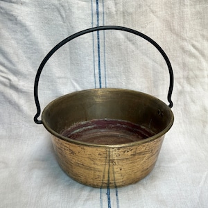 Puede incluir: Un caldero de latón vintage con un asa de metal negro. El caldero tiene forma redondeada y un aspecto desgastado y envejecido. El interior muestra signos de uso, con un tono rojizo cerca de la base. El asa se arquea sobre la parte superior.