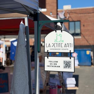 May include: A double-sided hanging sign with a dark green metal bracket. The top sign features a green soap icon and the text LA PETITE ARTI HANDMADE SOAPS. The bottom sign displays the text FOLLOW US ON INSTAGRAM next to a black and white QR code.