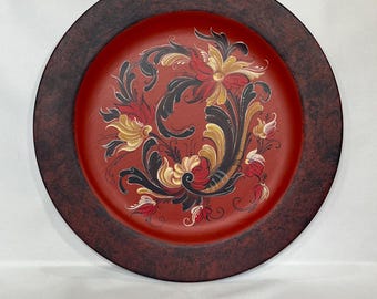 Op de afbeelding: Een ronde houten plaat met een donkerbruine rand en een rood midden. De plaat is versierd met een handgeschilderd bloemenontwerp in zwart, rood, geel en wit.