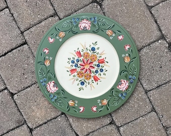 Op de afbeelding: Een ronde, handgeschilderde decoratieve schotel met een groene rand en een wit midden. De schotel heeft een bloemenontwerp met roze, oranje en blauwe bloemen en groene bladeren. De schotel ligt op een grijs bakstenen oppervlak.