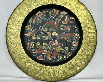 Op de afbeelding: Een ronde houten plaat met een zwarte achtergrond en een gouden rand. De plaat is versierd met een bloemmotief in rood, groen en geel.
