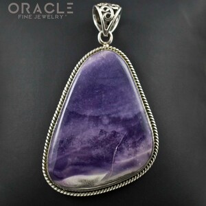 Puede incluir: Un colgante de plata con una piedra preciosa grande, de forma irregular, de color p&uacute;rpura y blanco, engastada en un bisel de plata con un borde de cuerda. El colgante tiene una anilla de plata con un dise&ntilde;o intrincado.