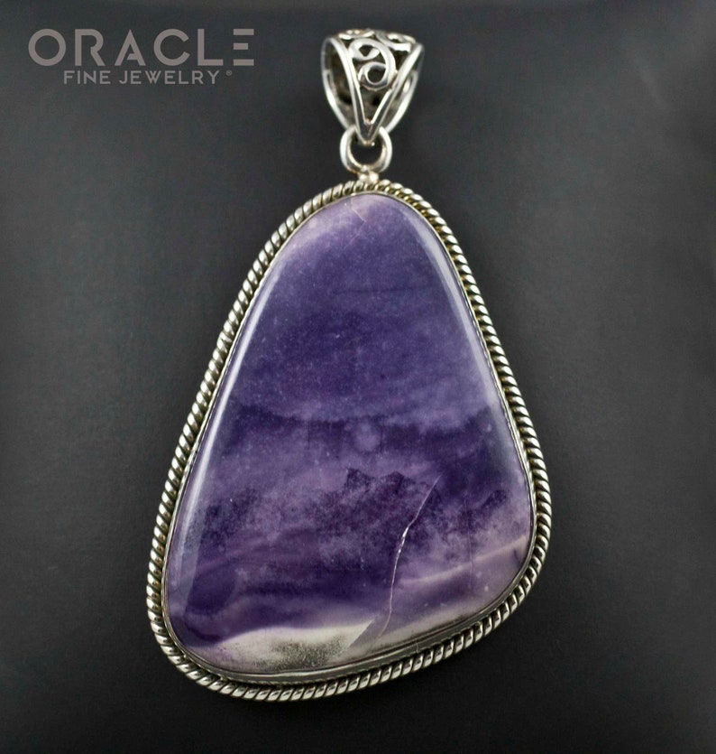 Puede incluir: Un colgante de plata con una piedra preciosa grande, de forma irregular, de color p&uacute;rpura y blanco, engastada en un bisel de plata con un borde de cuerda. El colgante tiene una anilla de plata con un dise&ntilde;o intrincado.