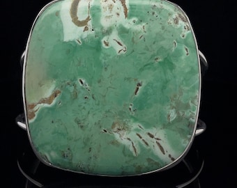 May include: A silver cuff bracelet featuring a large, square-shaped green turquoise stone. The stone has a mottled pattern of green, brown, and white.