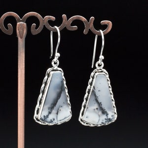 May include: A pair of silver dangle earrings featuring white and grey dendritic agate stones. The stones are set in a silver frame with a rope-like design.