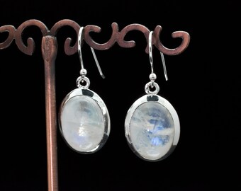 May include: A pair of oval-shaped moonstone earrings with a silver setting. The moonstones have a milky white appearance with subtle blue and iridescent flashes. The earrings are displayed on a decorative metal stand against a black background.