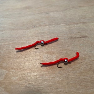 May include: Two red fishing lures shaped like worms, each with a silver bead and a small hook. The lures are lying on a light brown wooden surface. These artificial worms are designed for fishing.