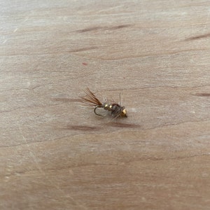 May include: A close-up of a small, artificial fishing fly. The fly has a gold-colored head, a dark hook, and brown and tan feather details. It is resting on a light brown wooden surface.