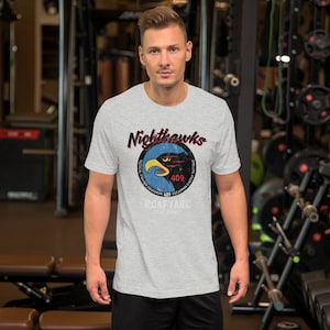 Könnte beinhalten: Ein graues T-Shirt mit einer Grafik im Vintage-Stil eines schwarzen Adlers mit dem Text "Nighthawks" und "409 Tactical Fighter Squadron" in einem Kreis. Der Text "RCNAF TARC" befindet sich unter dem Kreis.