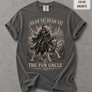 May include: A granite-colored t-shirt featuring a graphic print of a knight in armor riding a large wolf. The text reads HEAR YE! HEAR YE! THE FUN UNCLE HAS ARRIVED. The design includes a castle in the background and a forest setting.