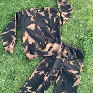 Puede incluir: Un conjunto de sudadera y pantalón de chándal de color negro y marrón con estampado tie-dye. La tela tiene un patrón tie-dye descolorido.