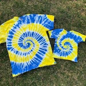 Może przedstawiać: Dwie koszulki tie-dye w niebiesko-żółtym wzorze spiralnym. Większa koszula jest w rozmiarze dla dorosłych z długim rękawem, a mniejsza koszula jest w rozmiarze dla dziecka z krótkim rękawem.