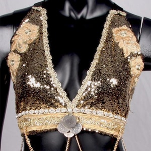 Puede incluir: Top halter dorado con lentejuelas y cuello en V. Presenta bordados dorados y una banda decorativa con monedas plateadas. Cadenas doradas cuelgan de la parte inferior. Se muestra en un maniquí negro. Adecuado para danza o espectáculos.