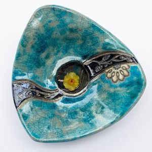 May include: A triangular ceramic bowl featuring a vibrant turquoise crackle glaze finish. The centre contains a small, raised yellow flower detail. Two dark, curved bands with intricate white floral patterns extend from the centre to the edges.