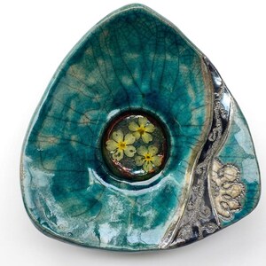 May include: A triangular ceramic bowl with a teal crackle glaze finish. The center features a circular inset containing three small, pressed yellow flowers. One side of the bowl has a dark, textured border with a white, lace-like floral pattern.