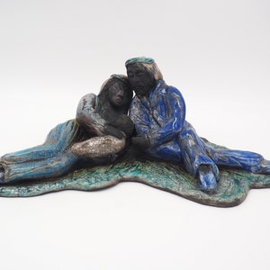 Pode incluir: Escultura de cerâmica de um casal sentado em uma base verde. A mulher está vestindo um vestido azul e branco e o homem está vestindo uma túnica azul.