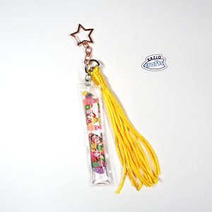May include: A clear keychain with a rectangular fruit-filled charm, a yellow tassel, and a star-shaped clasp. The charm contains colorful fruit slices. The keychain is attached to a metal ring.