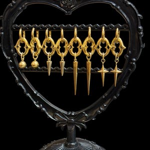 May include: A set of eight gold-toned hoop earrings hanging on a black heart-shaped display stand. Each hoop features a decorative link and a dangling charm, including two spheres, four spikes of varying lengths, and two four-pointed star shapes.