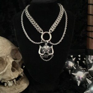 May include: A silver chain necklace with a large silver pendant featuring a snarling animal head. The necklace is layered with two other silver chains, one with a large ring.