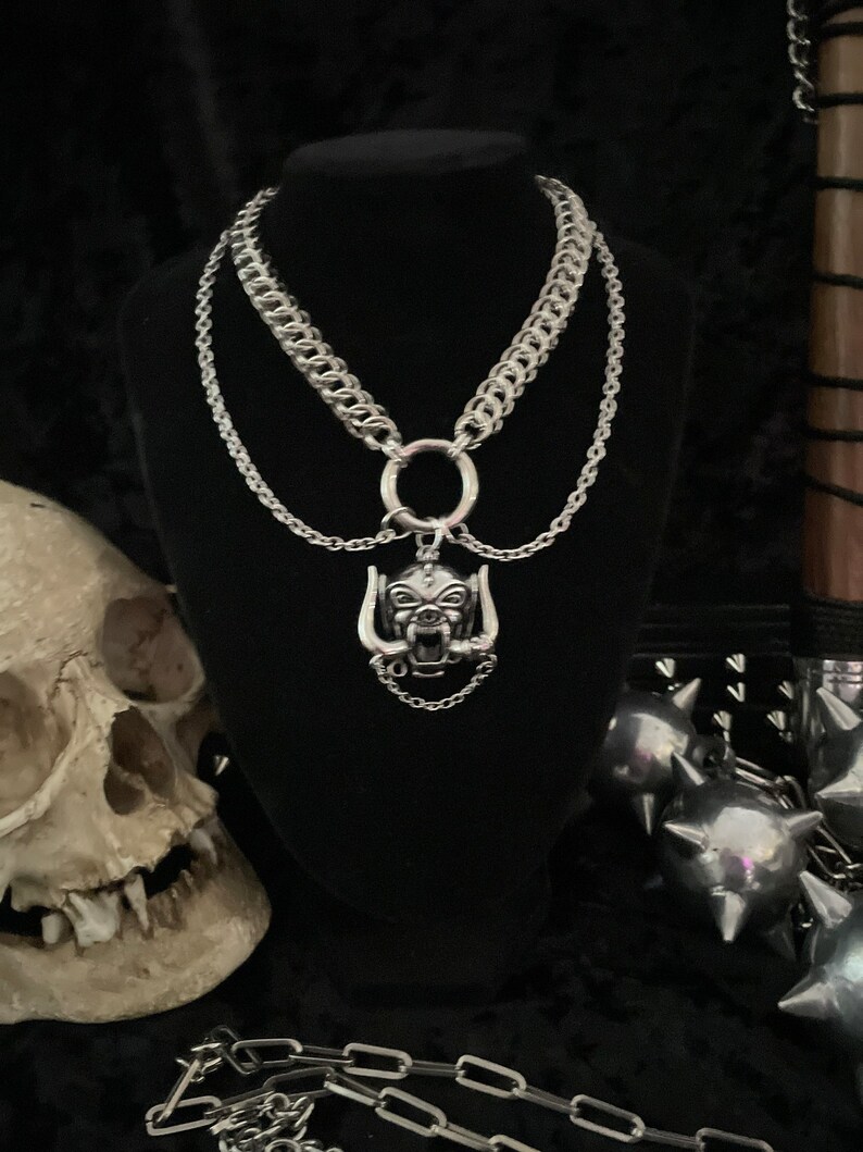 May include: A silver chain necklace with a large silver pendant featuring a snarling animal head. The necklace is layered with two other silver chains, one with a large ring.