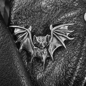 Peut inclure: Une épingle en forme de chauve-souris argentée avec des ailes détaillées et une tête de chauve-souris réaliste. L'épingle est fixée à une veste en cuir noire.